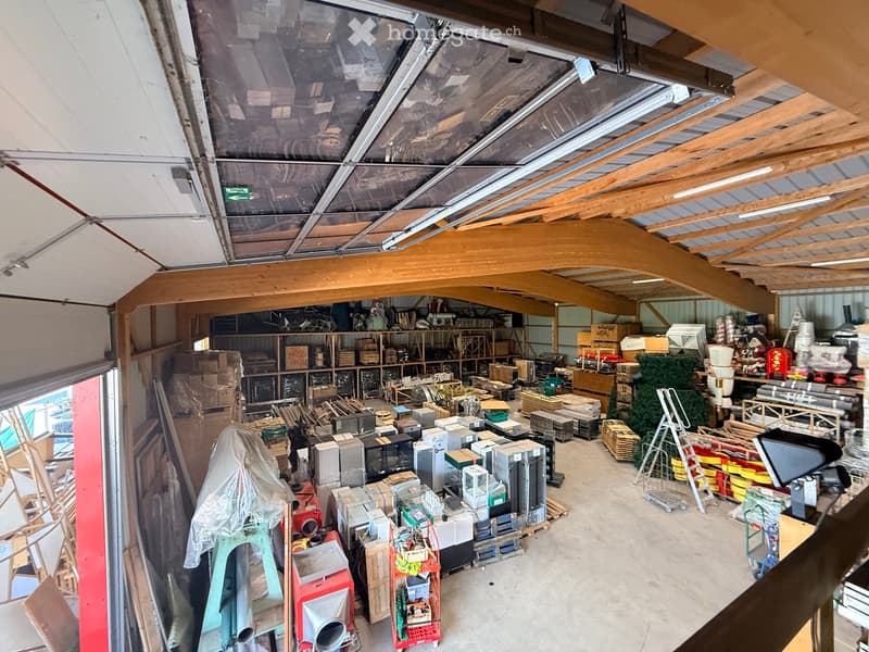 A spacious, high-ceilinged storage area with exposed wooden beams and a translucent roof section. The space is filled with various items including shelves, boxes, appliances, tools, and a ladder, organized in a functional layout.