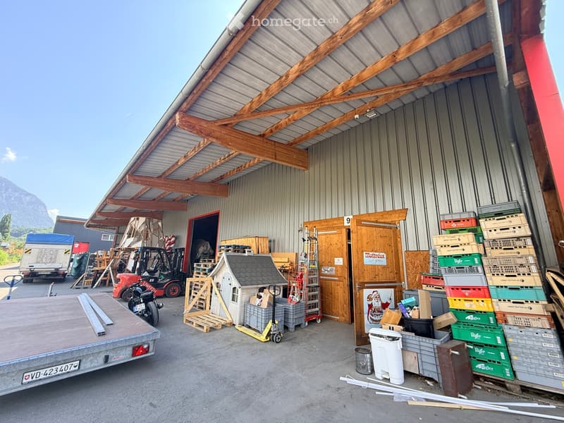 Warehouse or storage area with a metal roof and wooden beams, containing various items such as crates, pallets, a small playhouse, a forklift, and a trailer.