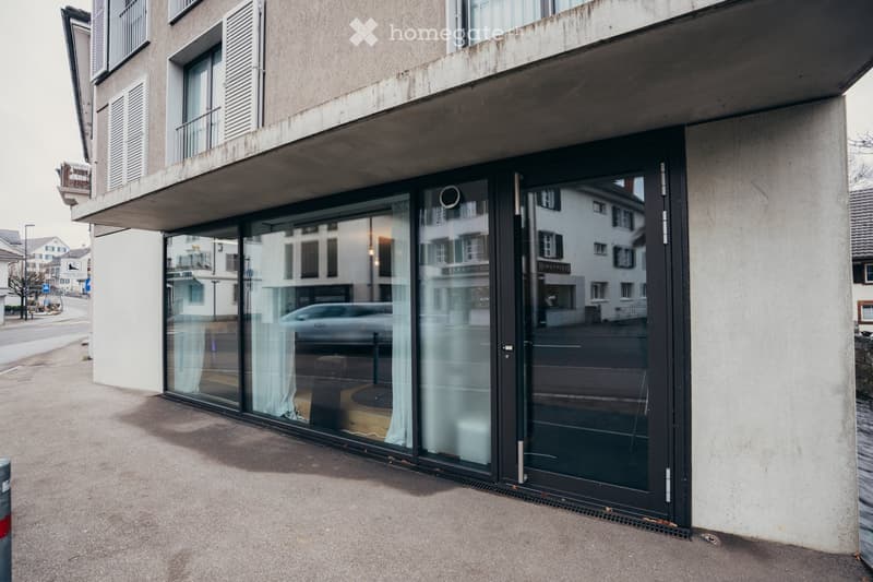 Modern building with gray concrete facade, glass windows, balcony with shutters, entrance door with large glass panels.