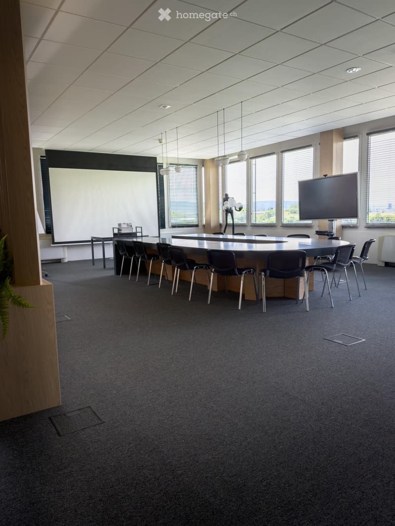 Modern conference room with a large round table, chairs, two screens, and large windows with blinds.