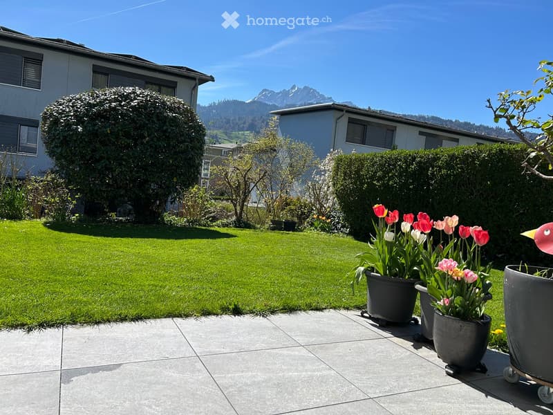 Well-maintained garden with a green lawn, trimmed hedges, and potted tulips on a tiled patio, with a scenic mountain view in the background.