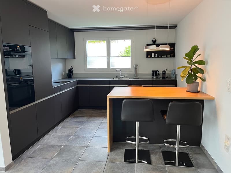 Modern kitchen with dark grey cabinets, a central wooden island with two black stools, and various appliances including an oven, sink, and fridge. There is a potted plant on the island and pendant lights hanging above.
