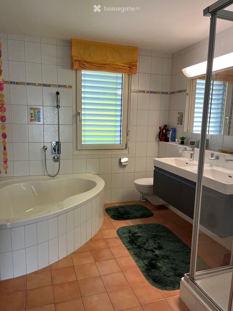 Modern bathroom with a bathtub, toilet, double sink, mirror, and showerhead. Features tiled walls, a window with blinds, and two green rugs on the floor.