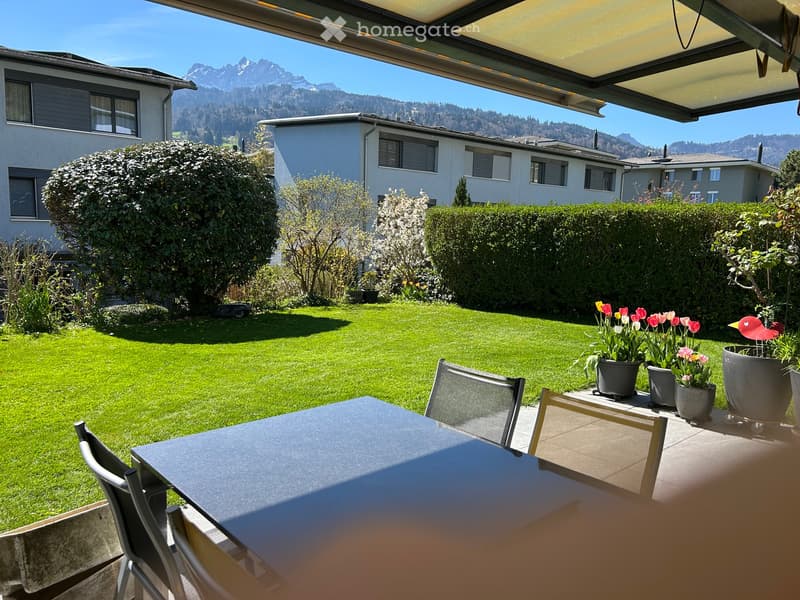 Well-maintained garden with a patio area featuring a table, chairs, and flower pots, surrounded by neatly trimmed hedges and a lawn, with a scenic mountain view in the background.