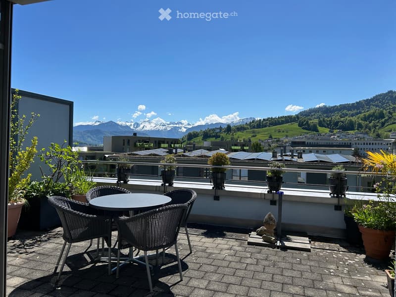Balcony with a round table and four wicker chairs, surrounded by potted plants, overlooking a scenic mountain view.