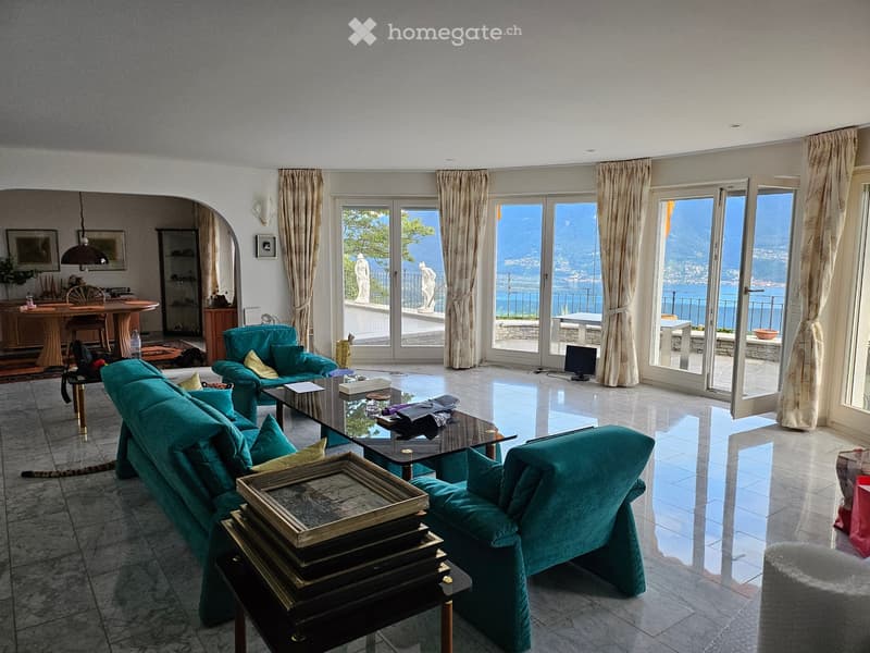 large windows with curtains, balcony, two blue chairs, round coffee table, large sofa, carpet, paintings, picture frames, table with objects
