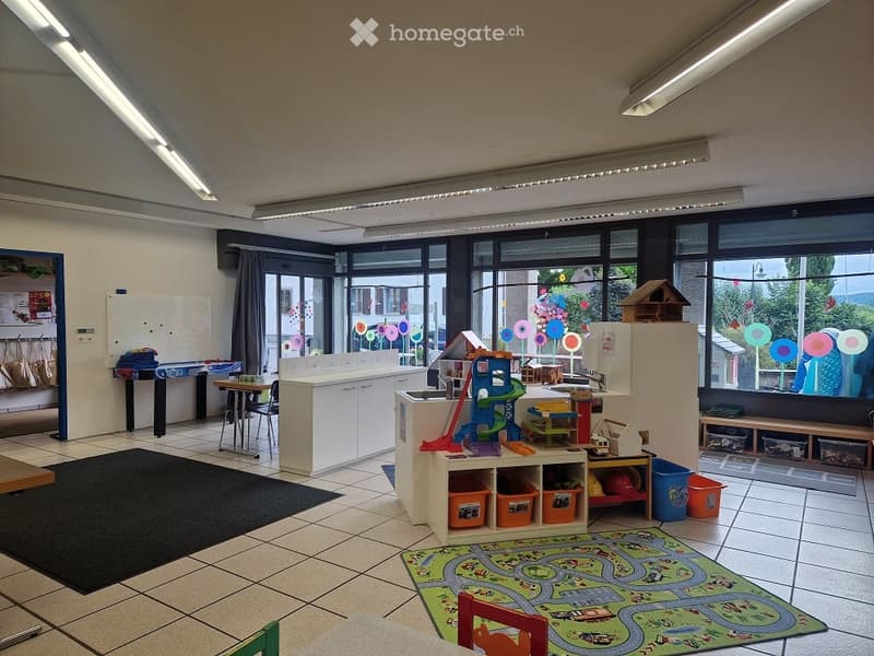 Bright, open room with large windows, tiled floor, and various children's furniture and toys including a playmat, shelves, and a small table.