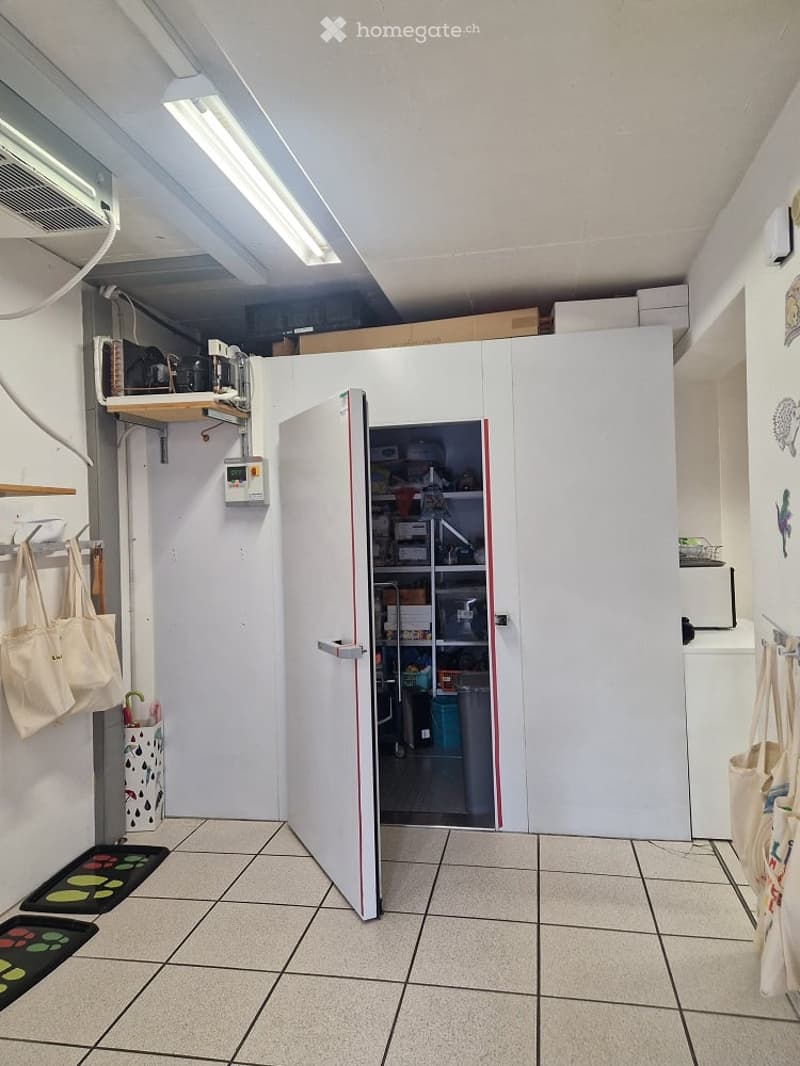 A small utility or storage room with a large white refrigerator, shelves filled with items, and several hanging bags. The room has tiled flooring and fluorescent lighting.