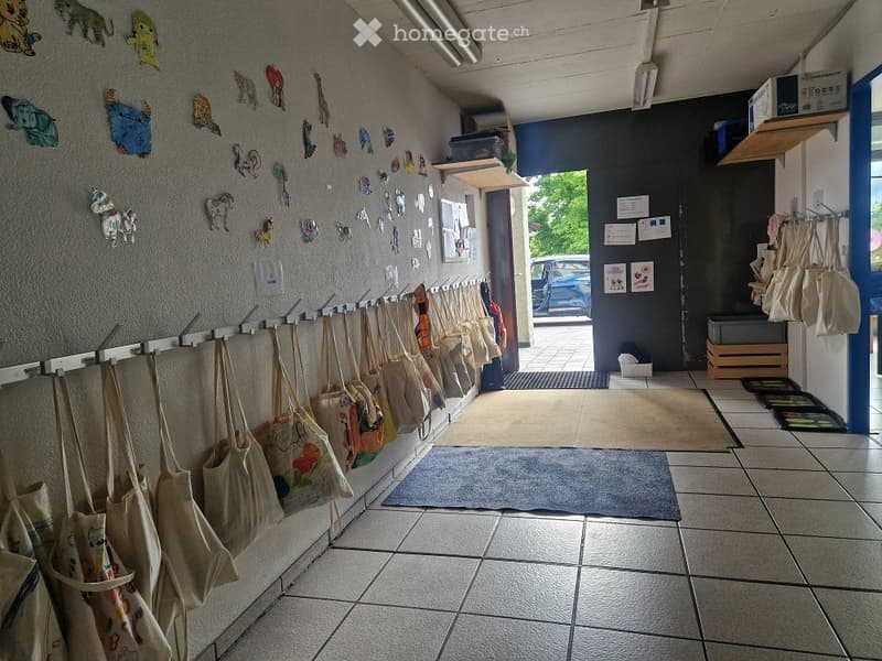 A hallway with tiled flooring, a row of hooks holding bags, and colorful artwork on the walls. A door leads outside, and there are additional storage items and a small rug in the foreground.