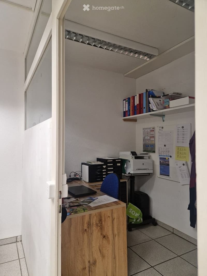 A small, well-lit office space with a wooden desk, a blue chair, a printer, a laptop, shelves with binders, and a calendar on the wall.