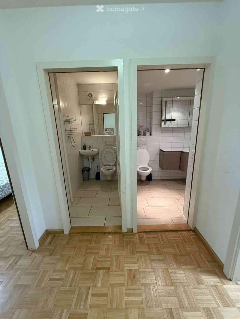 Hallway with wooden flooring leading to two bathrooms, one with a toilet and sink, the other with a toilet and cabinet.