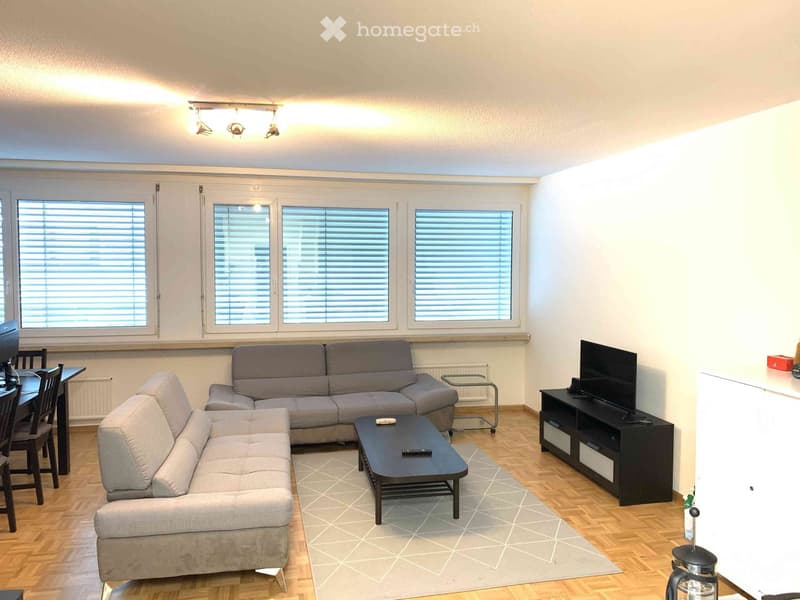 A modern living room with light-colored walls, wooden flooring, and large windows with blinds. Features include a grey sectional sofa, a black coffee table, a TV stand with a flat-screen TV, and a dining area with a table and chairs.