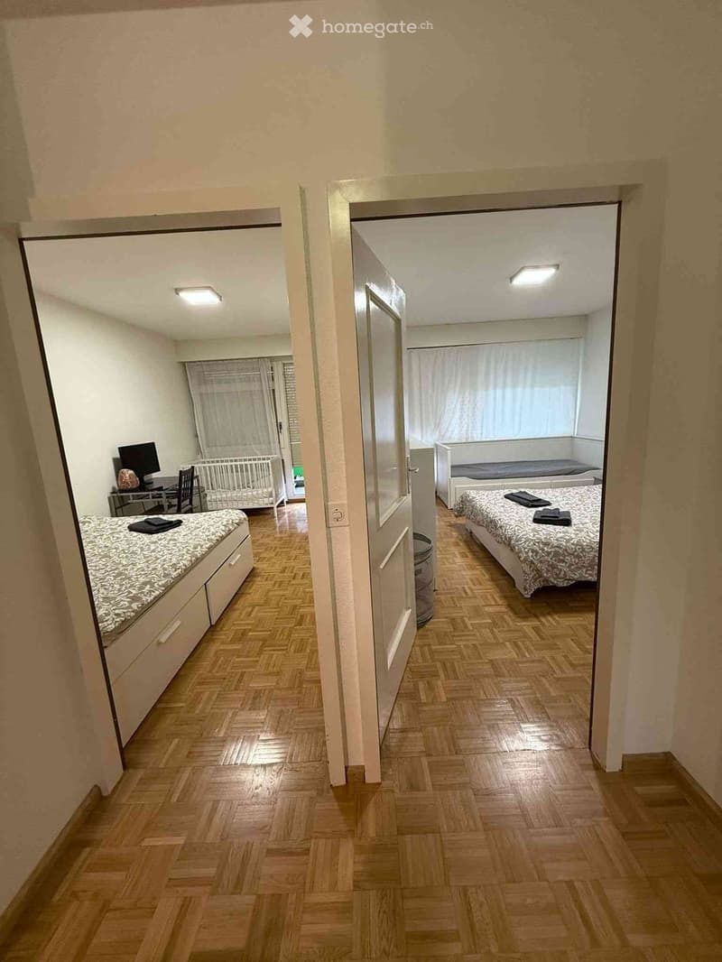 A hallway leads to two furnished rooms with beds, a desk, and a baby crib, featuring wooden flooring and white walls.