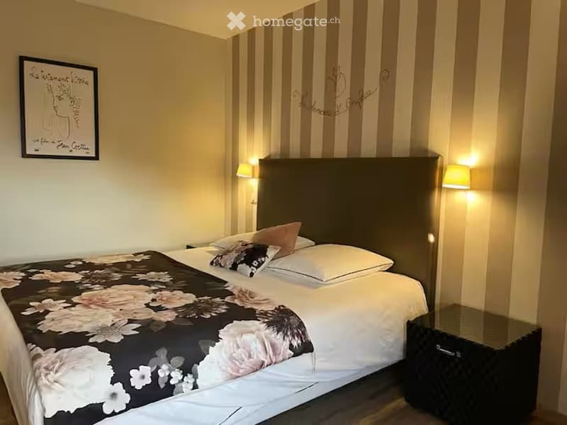 Cozy bedroom with a large bed, decorative pillows, a nightstand, and wall art. The room features warm lighting and striped wallpaper.