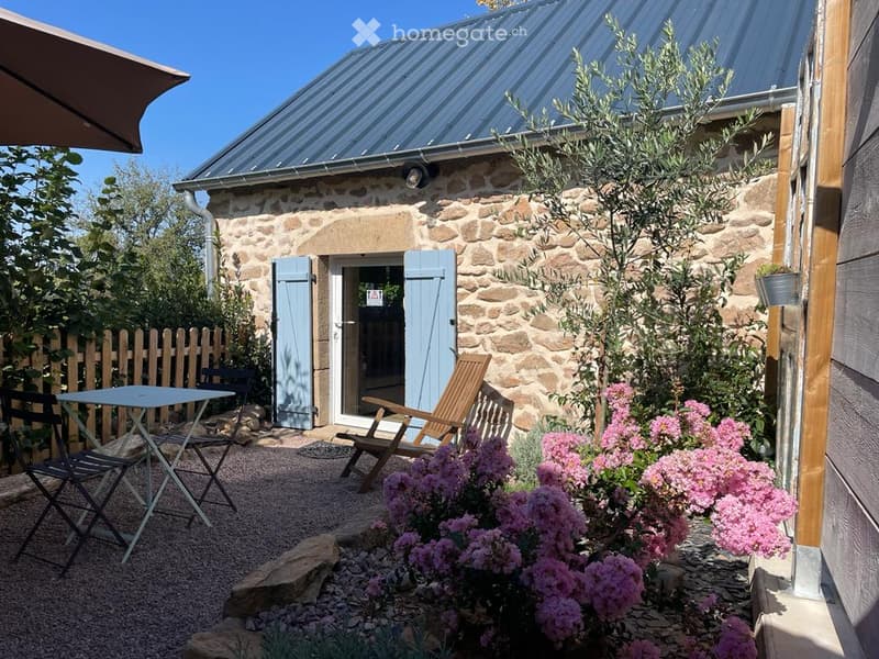 A small outdoor seating area with a table, chairs, and a wooden deck chair, surrounded by pink flowering bushes and a stone building with a metal roof and blue shutters.
