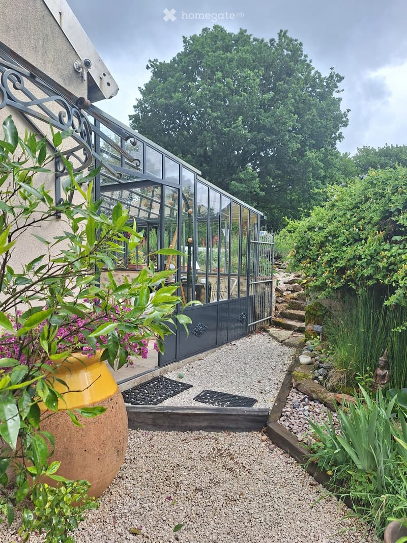 Garden area with a greenhouse, gravel pathway, large ceramic pot with flowers, and lush greenery.
