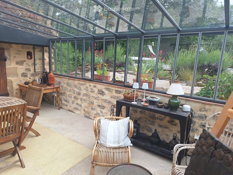 A cozy, well-furnished greenhouse with large glass windows, stone walls, and a variety of decorative items including lamps, tables, and chairs.