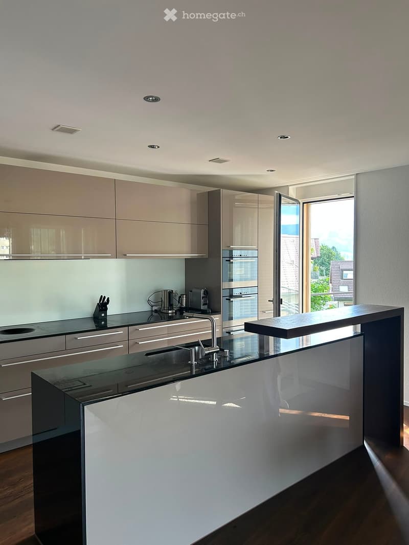 Modern kitchen with sleek, light-colored cabinets, a black countertop, and stainless steel appliances, including a sink, faucet, coffee maker, and oven. The space features a central island with a glass partition and ample natural light from large windows.