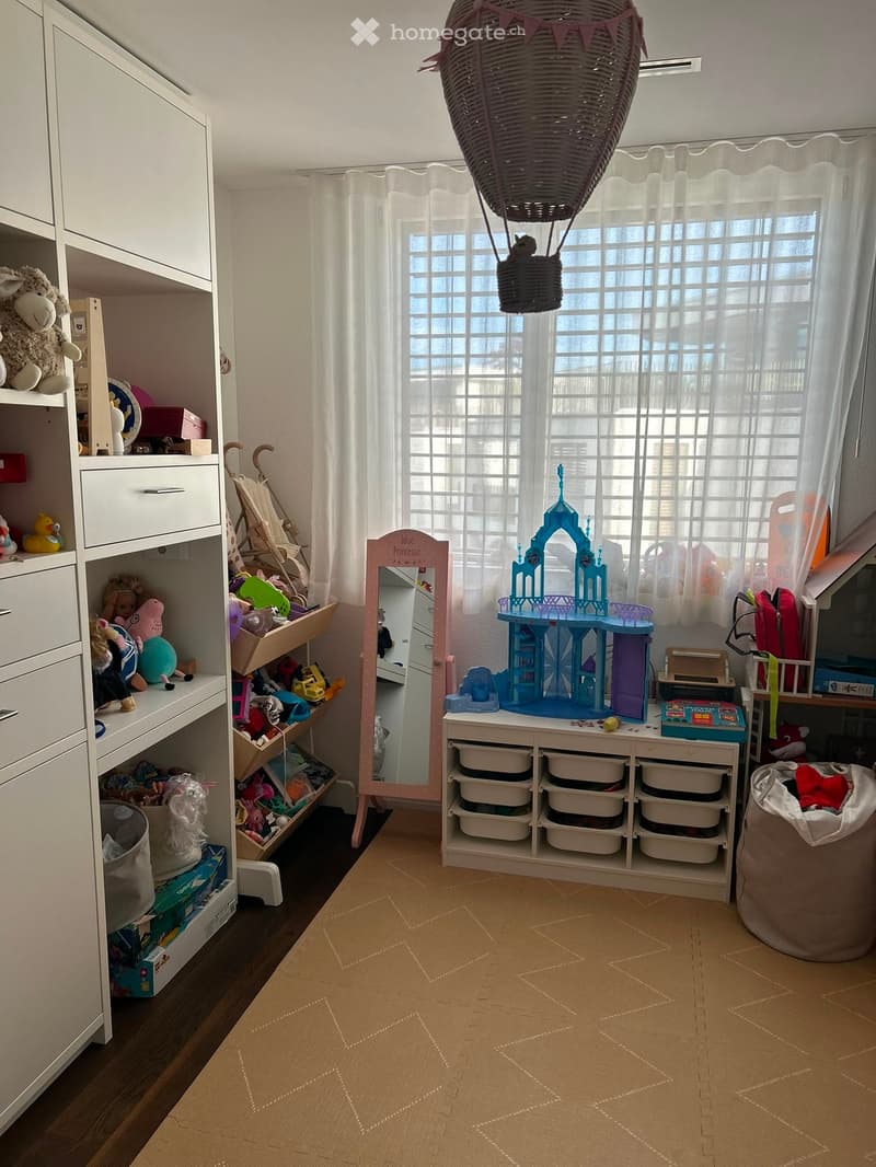 A well-lit children's room with a large window, a toy castle on a shelf, storage units, a mirror, and various toys scattered around.