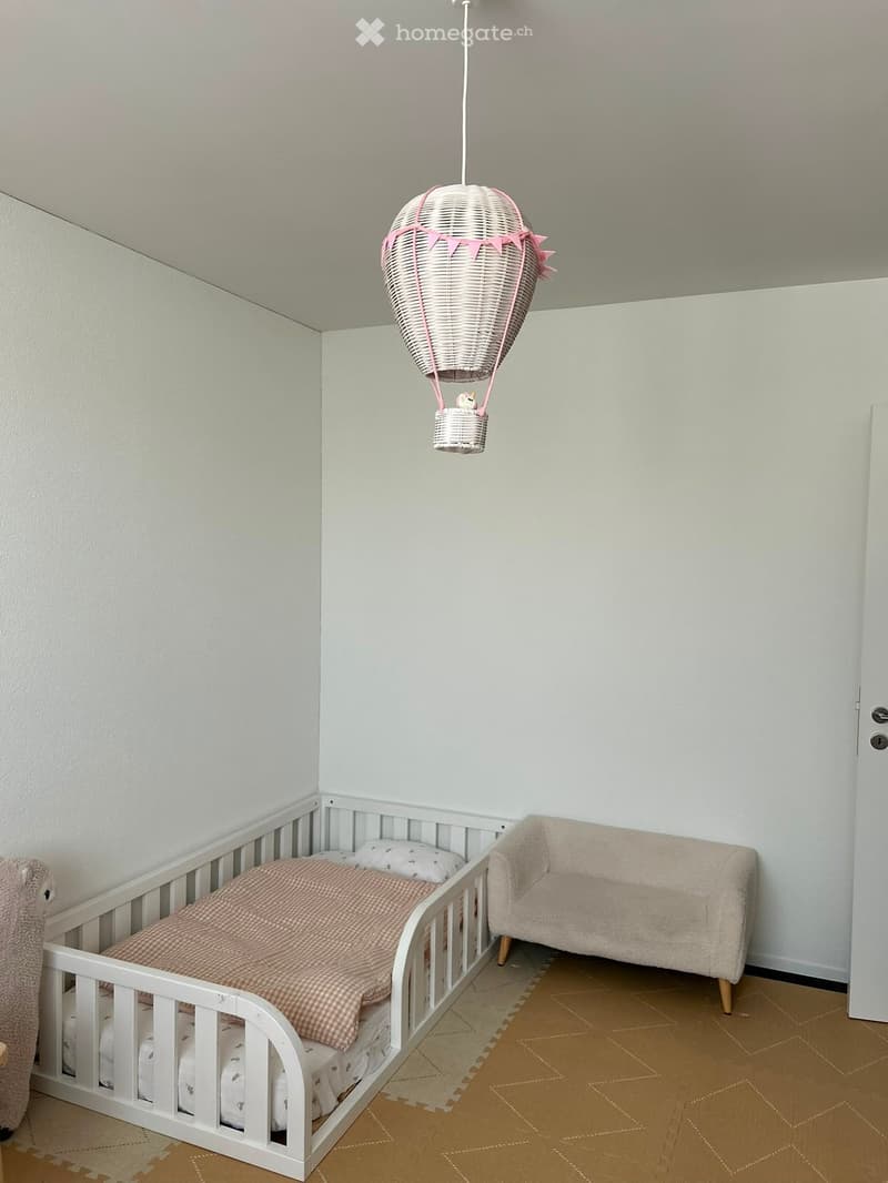 A cozy children's bedroom with a white crib, a small sofa, a hanging balloon decoration, and a plush toy on the floor.