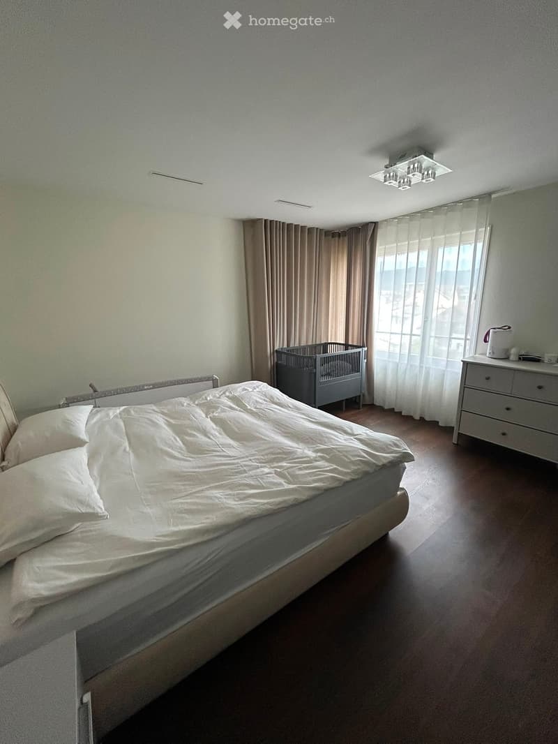 A well-lit bedroom with a large bed, a baby crib, a dresser, and curtains on the windows.