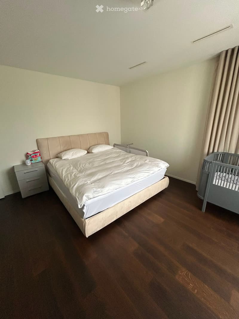 A well-lit bedroom with a large bed, a nightstand, a crib, and a curtain on the window. The room features wooden flooring and neutral-colored walls.