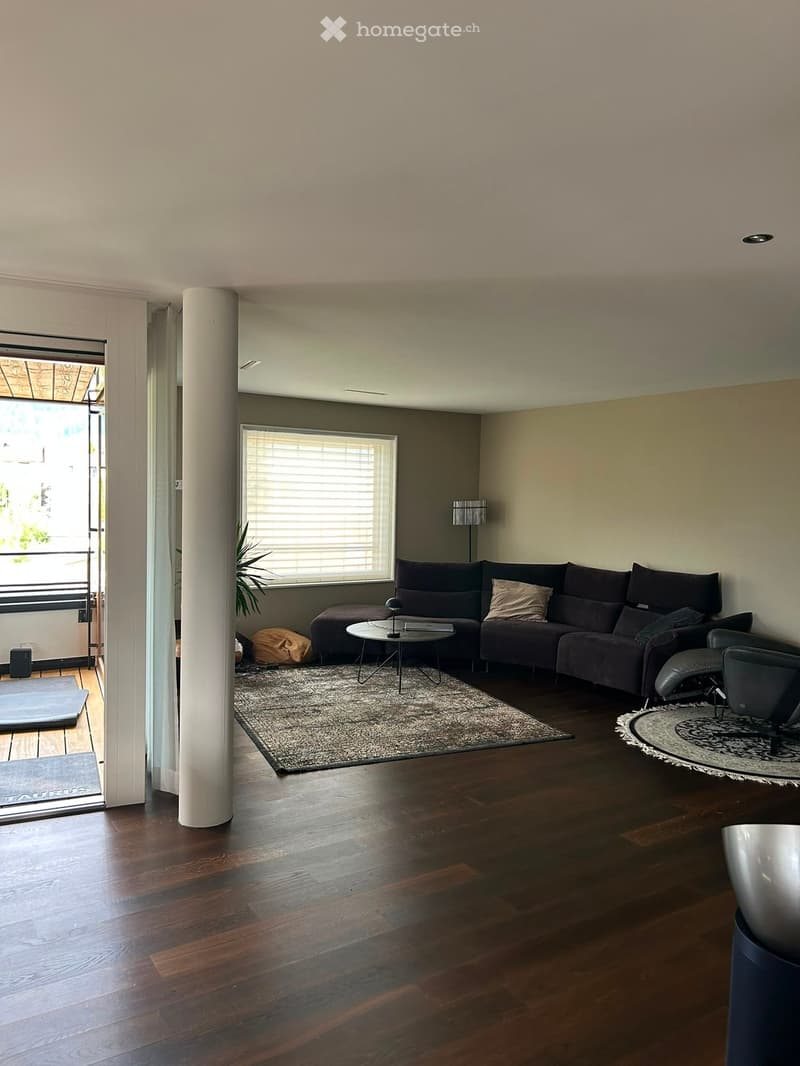 Modern living room with dark wood flooring, a sectional sofa, coffee table, and a large window with blinds. Features a tall white column and a view of a balcony area.