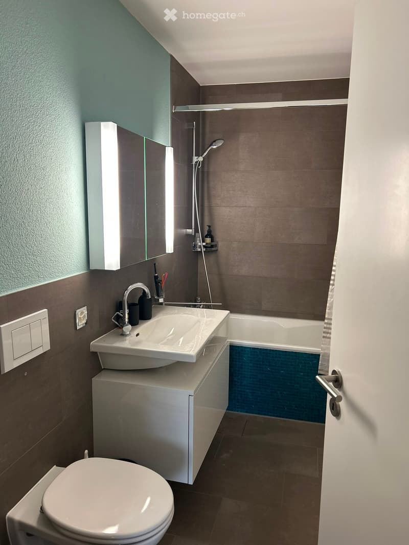 Modern bathroom with a white sink, toilet, bathtub, and showerhead. Features a large mirror with built-in lighting, a sleek faucet, and a dark brown tiled wall.