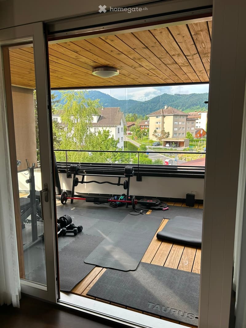 Balcony with wooden floor, exercise equipment including a bench, weights, and yoga mats, overlooking a scenic town with mountains in the background.