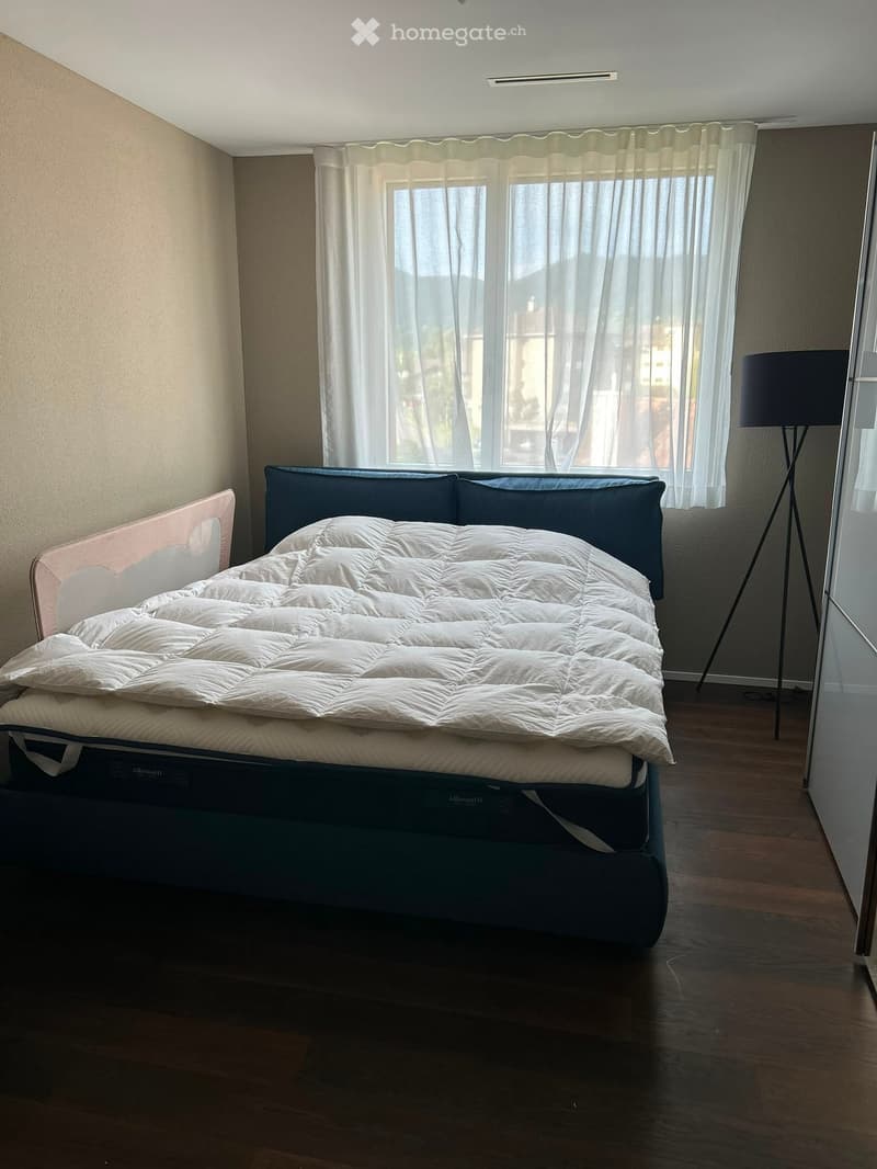 Modern bedroom with a large bed, white duvet, dark blue headboard, and a floor lamp near the window with sheer curtains.