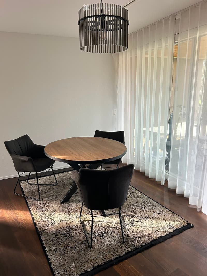 Modern dining area with a round wooden table, four black chairs, a patterned rug, and a contemporary chandelier.