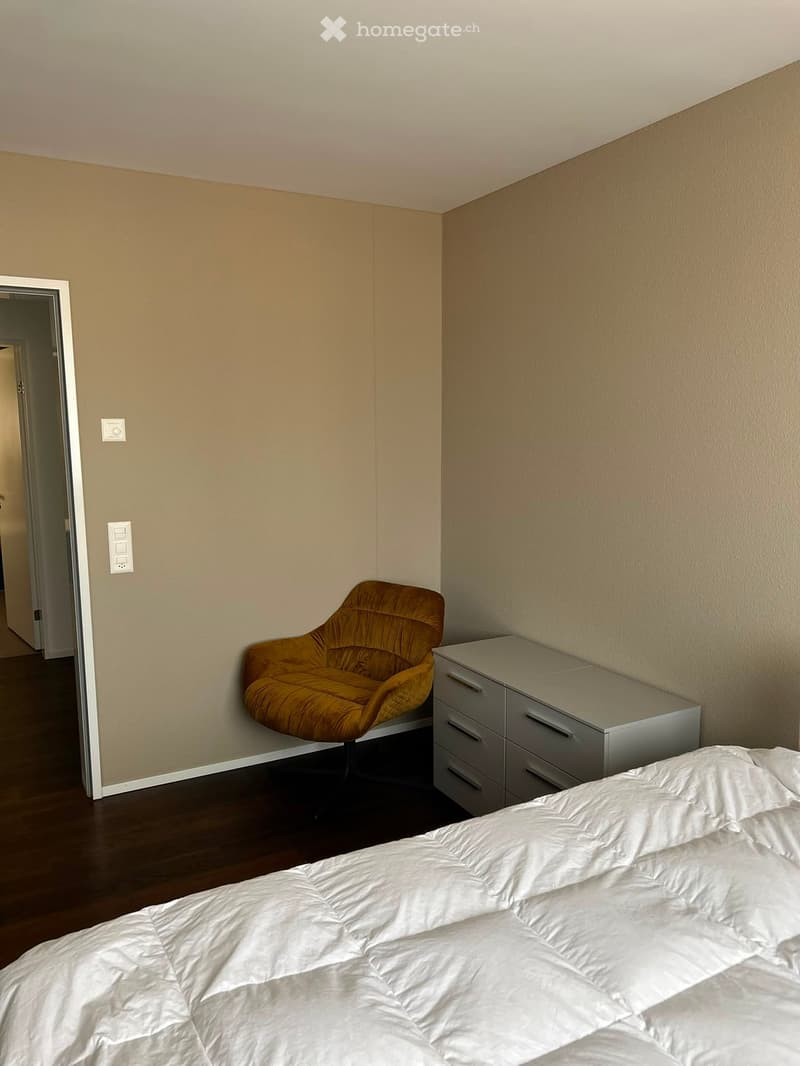 A modern bedroom with a neatly made bed, a sleek nightstand, and a stylish orange armchair. The room features light-colored walls and a dark wooden floor.