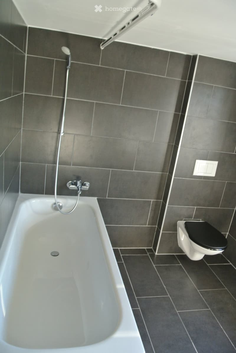 Modern bathroom with a white bathtub, grey tiles, and a toilet with a black seat.