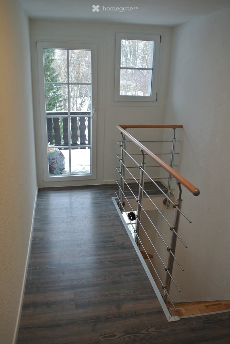 A modern hallway with wooden flooring, white walls, and a staircase with a metal railing and wooden handrail. Two windows provide natural light, and a door leads to an outdoor area.