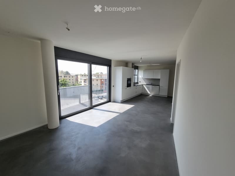 Empty room with grey carpeted floor, white walls, large sliding glass door leading to a balcony, and a small kitchen area with white cabinets and appliances visible in the background.