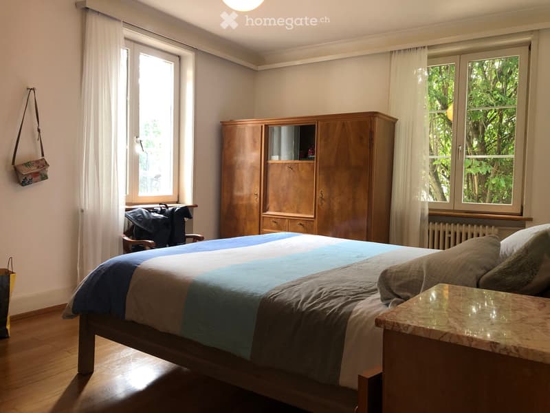 Cozy bedroom with wooden bed, blue and grey bedding, large wooden wardrobe, nightstand, and a chair near the window.