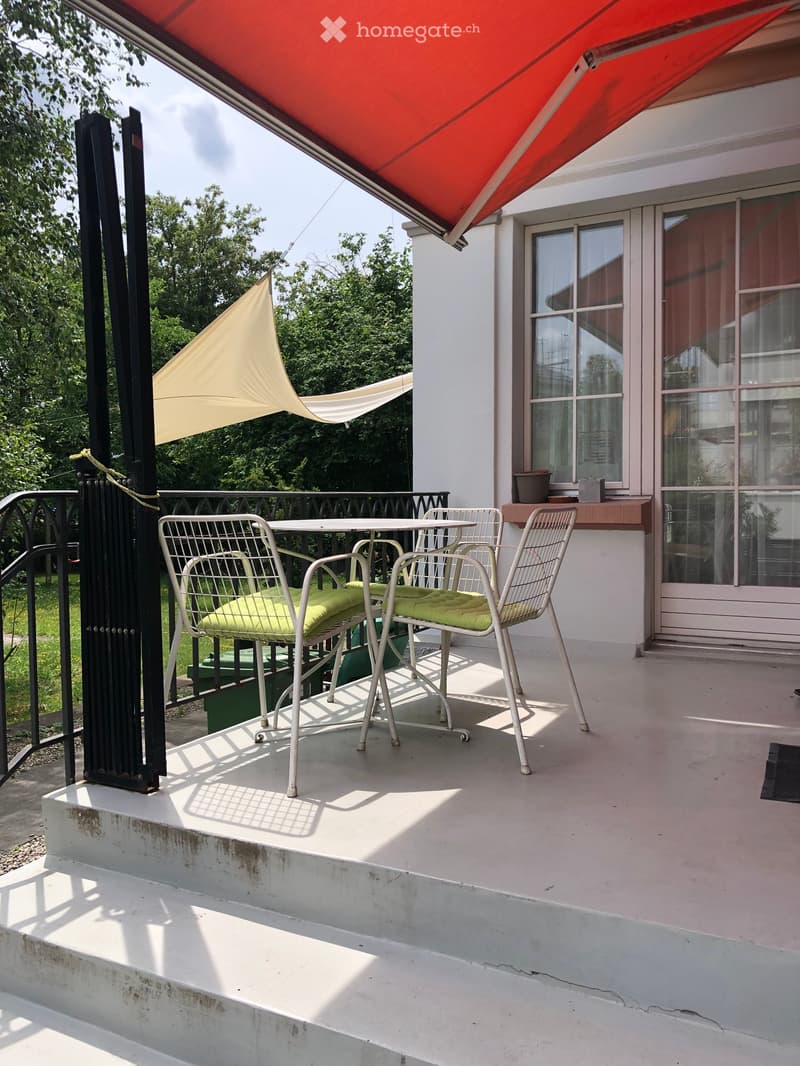 Balcony with a table and four chairs, a red awning, and a view of greenery.