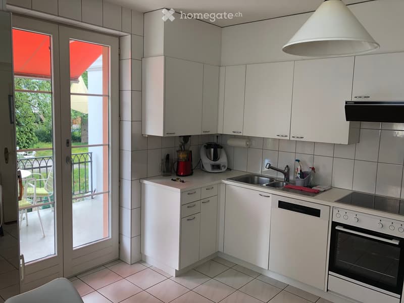 Modern kitchen with white cabinets, stainless steel sink, oven, dishwasher, and various appliances. A large window opens to a balcony with outdoor furniture visible.