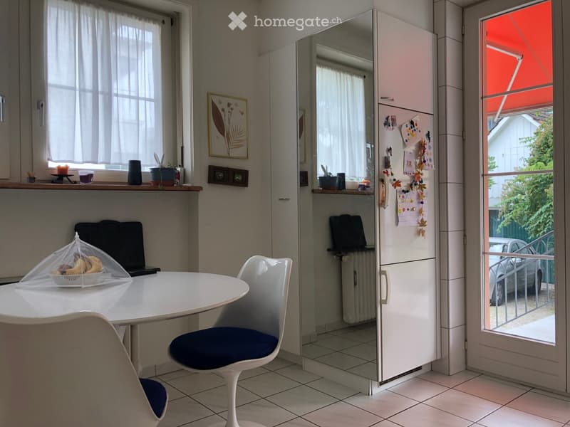 A modern, well-lit room with a round white table, two white chairs with blue cushions, a laptop, a refrigerator, a mirror, and a framed picture on the wall. The room has tiled flooring and large windows with sheer curtains.