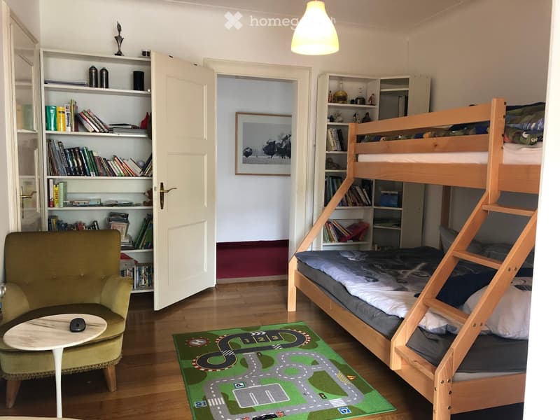 A cozy bedroom with a bunk bed, a green armchair, a small table, and built-in bookshelves. The room features a race track rug and a framed picture on the wall.