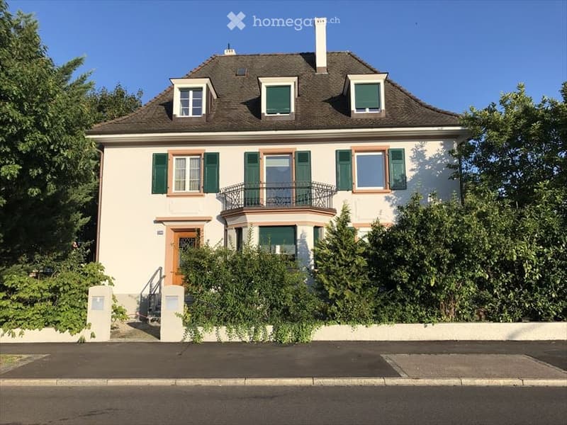 Two-story house with white walls, green shutters, and a dark roof. Features a balcony with a metal railing and a small garden in front.