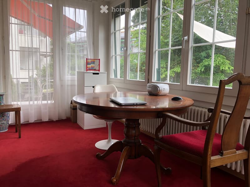 A furnished room with a round wooden table, a wooden chair with red upholstery, a laptop, a small radio, a radiator, a small stool, and a decorative vase near the window.