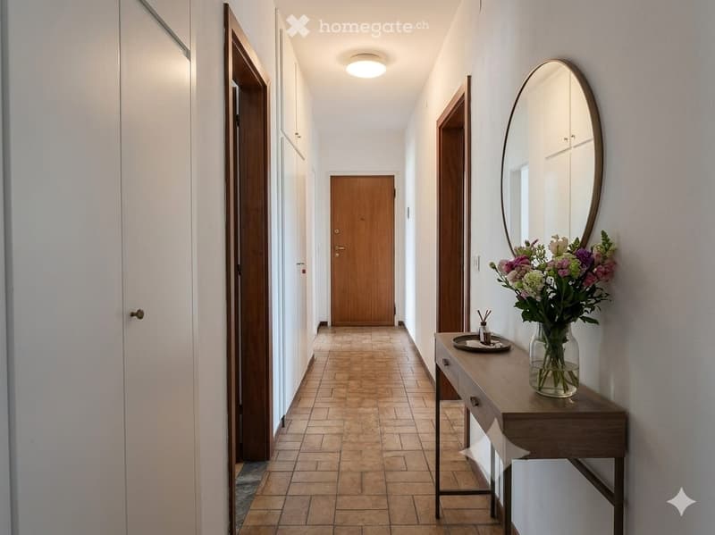 A modern hallway with tiled flooring, white walls, and a round mirror on the right side. A small console table holds a vase of flowers and a tray with candles.