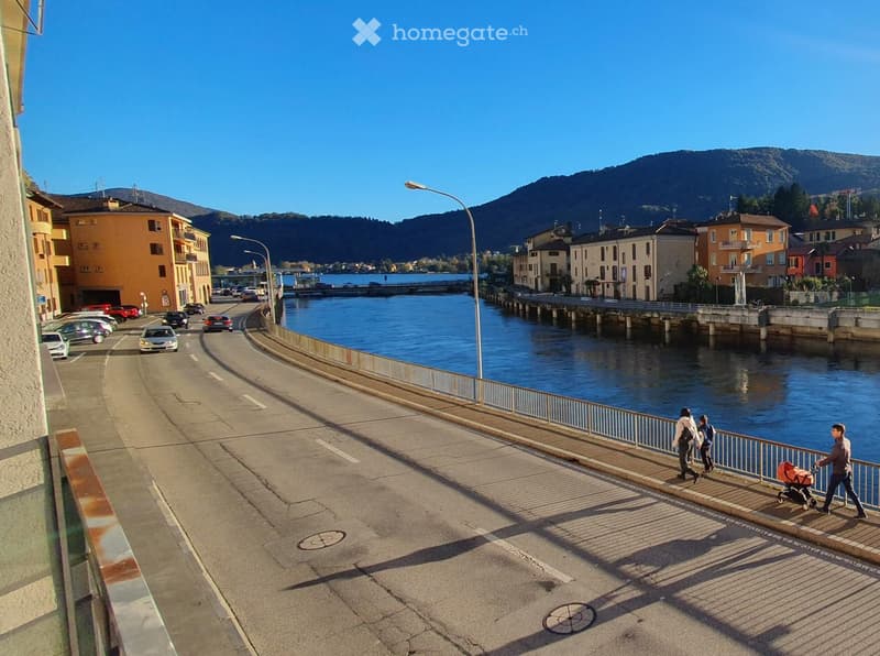 A road with a river on one side, a metal railing, street lights, people walking on the sidewalk, buildings, mountains in the distance, cars parked on the side of the road.