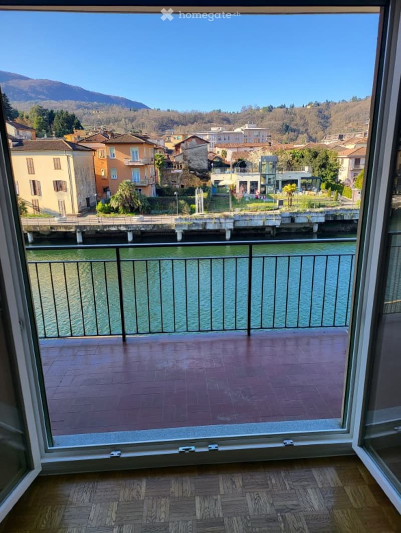 Open sliding glass door, balcony with metal railing, tiled floor, view of a river and buildings