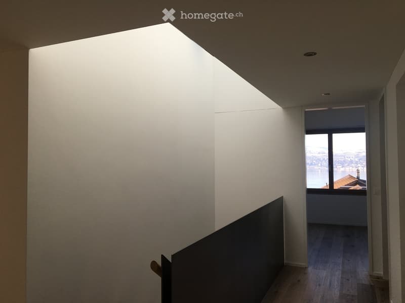 Modern hallway with white walls, wooden flooring, and a large skylight. A doorway leads to a room with a view of the water and cityscape.