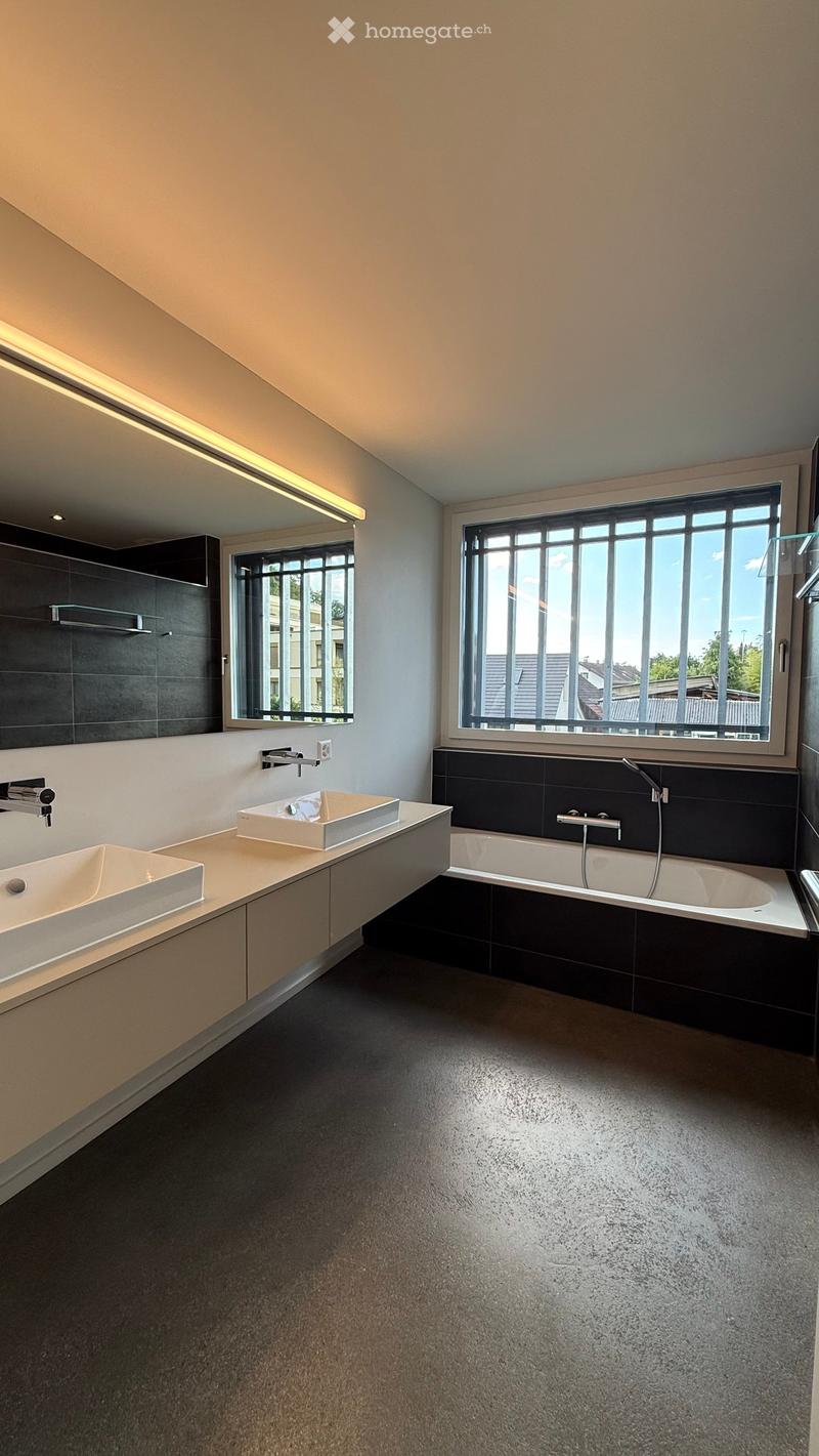 Modern bathroom with two sinks, a bathtub, and large windows providing natural light. Features sleek, contemporary design with dark tiles and light countertops.