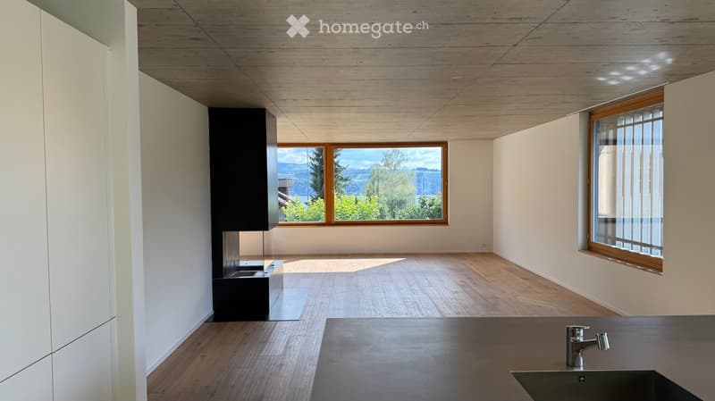 Modern, minimalist kitchen with light wooden flooring, white walls, and large windows offering a scenic view. Features include a sleek black cabinet, a built-in sink with a modern faucet, and a recessed ceiling light.