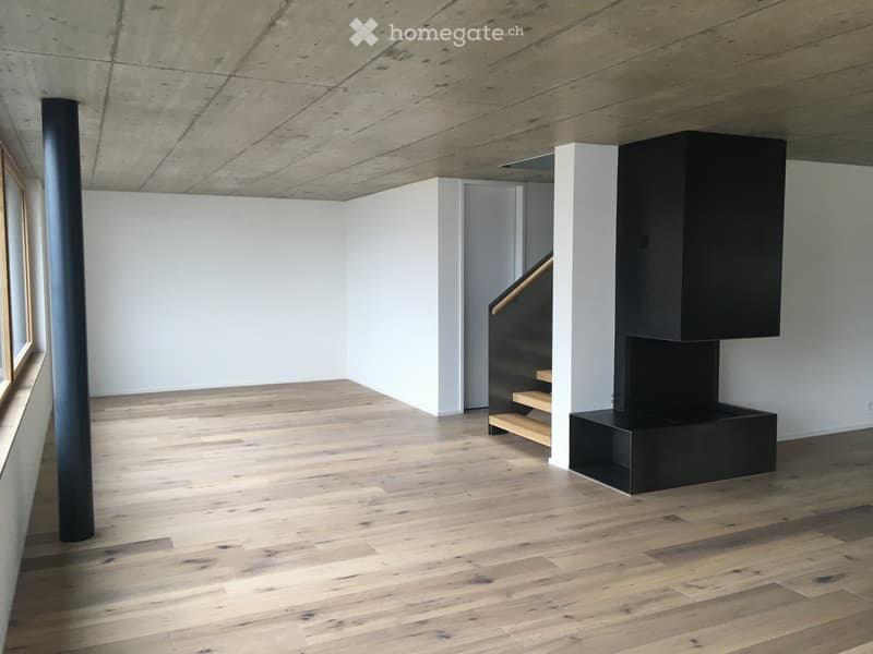 Empty room with light wooden flooring, white walls, and a black staircase leading upwards.