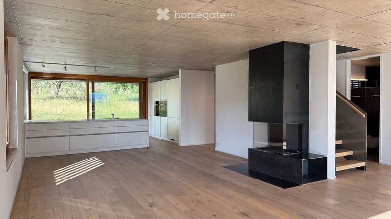 Modern, open-plan living and dining area with light wooden flooring, white kitchen cabinets, a sleek black fireplace, and a staircase leading to an upper level.
